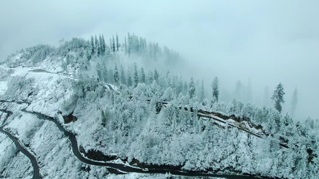 4K Aerial shot of breathtaking winter landscape with winding road on mountain at Manali, Himachal Pradesh, India. Clouds covering pine trees on mountain peak during snow storm. Scenic landscape view.