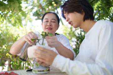 Asian Mature woman and teenage boy fixing soil and watering plants at garden, enjoying outdoor moments and learning plant care together.
