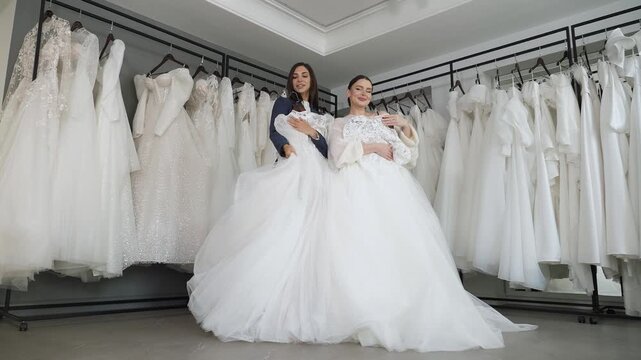 Two graceful young women, dressed in wedding attire, stand in a wedding salon, gazing into the camera and smiling. Behind them, a plethora of wedding dresses hangs on racks.