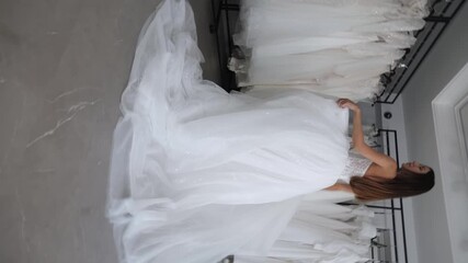 A gorgeous slender bride with long hair is spinning in a wedding dress in a bridal salon. The woman tries it on. Stable rotation of the camera in a circle. There are many wedding dresses in the salon.
