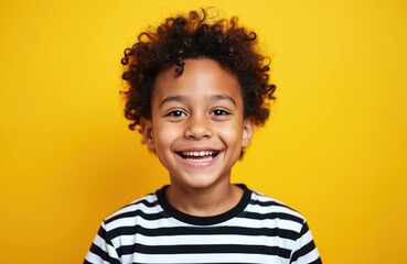 Small boy with curly dark hair smiles widely in a striped shirt against a bright yellow background. He has a joyful expression and looks directly at the camera.