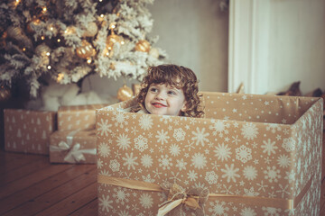 H&uuml;bsches kleines M&auml;dchen mit Locken sitzt in einem Geschenkkarton vor dem Weihnachtsbaum zu Weihnachten, Abendstimmung mit Lichtern Var. 4