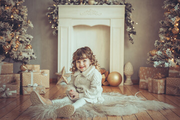 H&uuml;bsches kleines M&auml;dchen mit Locken sitzt vor einem Kamin mit Weihnachtsbaum zu Weihnachten, Abendstimmung mit Lichtern Var. 4