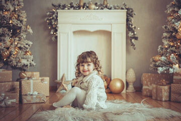 H&uuml;bsches kleines M&auml;dchen mit Locken sitzt vor einem Kamin mit Weihnachtsbaum zu Weihnachten, Abendstimmung mit Lichtern Var. 1