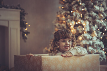 H&uuml;bsches kleines M&auml;dchen mit Locken sitzt in einem Geschenkkarton vor dem Weihnachtsbaum zu Weihnachten, Abendstimmung mit Lichtern Var. 1