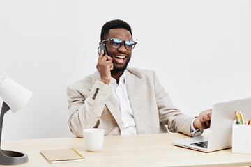 Smiling man wearing glasses talking on mobile phone while working at desk in modern office interior with laptop and stationery, cheerful professional male, business communication, casual style