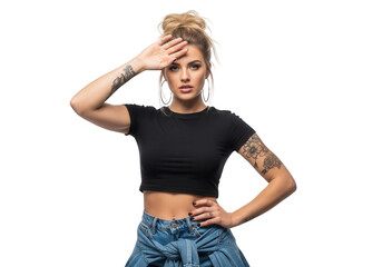 Young tattooed woman with blonde hair bun in black crop top and denim, hand on forehead, looking ahead with serious expression