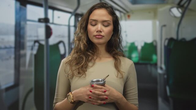 Young hispanic woman enjoys a mate beverage indoors on a bus, holding an infusion cup while savoring the moment, capturing a serene travel experience.