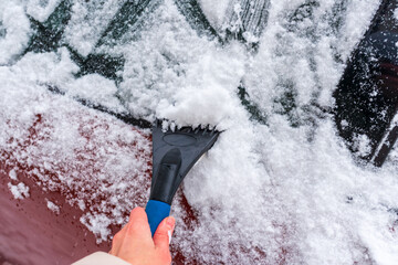 A hand uses a blue brush and scraper to remove fresh snow from a car windshield. Ice fragments and...