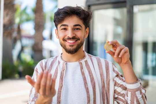 Handsome Arab man holding a Bitcoin at outdoors inviting to come with hand. Happy that you came
