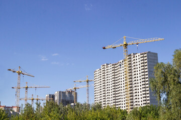 A building under construction and construction cranes among the trees. Blue sky