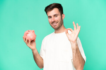 Young caucasian man holding piggybank isolated on green background showing ok sign with fingers