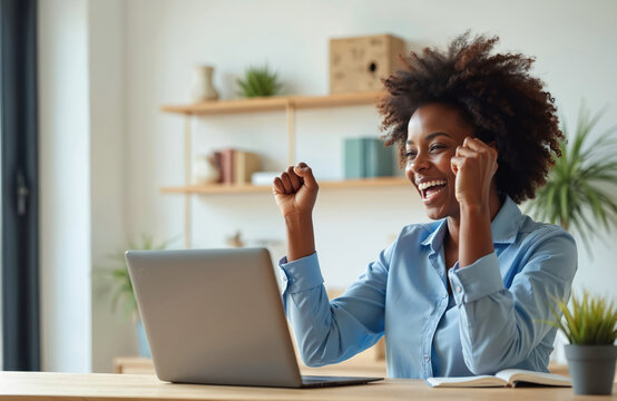 African woman rejoices at laptop screen. She celebrates success with raised fists and wide smile. Young professional works remotely from home office, achieving career goal. - Powered by Adobe