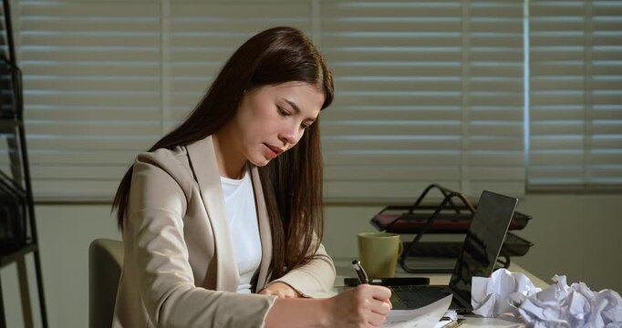 Asian businesswoman writing report at night in office, surrounded by crumpled paper, showing effort to correct mistakes and complete project, concept of determination, stress, and late work dedication