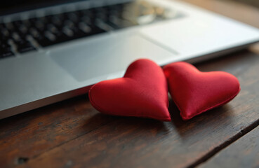 Two red heart shaped pillows lie on a wooden table next to a laptop computer. Hearts are on a brown wooden desk near a silver laptop keyboard. Valentine day love concept.