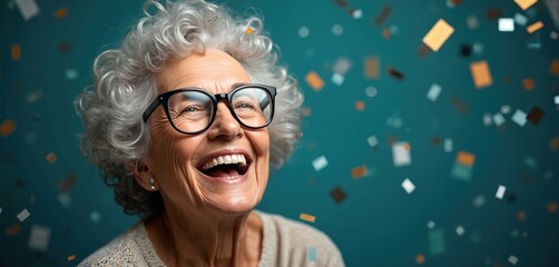 Happy senior woman laughs joyfully, wearing glasses with gray curly hair. Looks up with big smile as colorful confetti falls around on vibrant blue background. Celebrates special event personal