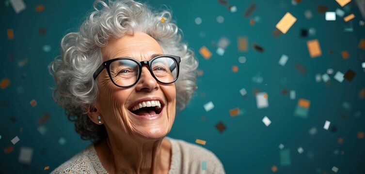Happy senior woman laughs joyfully, wearing glasses with gray curly hair. Looks up with big smile as colorful confetti falls around on vibrant blue background. Celebrates special event personal