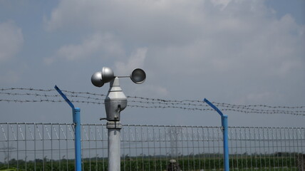 Obraz premium Anemometer measures wind speed at weather station near barbed wire fence on partly cloudy day, showcasing technology and environmental monitoring.
