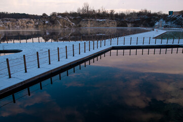Naklejka premium Snow-covered walkway over a calm lake with reflections of clouds.