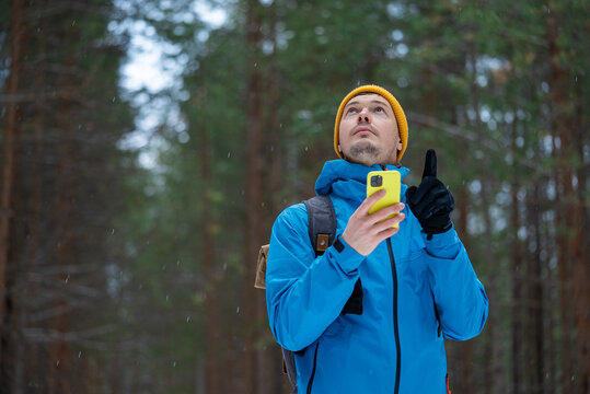 Man in a winter hat and jacket holding a bright yellow smartphone and gazing upwards, seeking direction and attempting to orient himself while being lost among the tall pine trees in a snowy forest