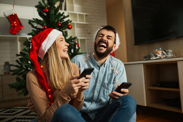 Two young people wearing Christmas hats, sitting together in a festively decorated apartment, using their smartphones while surrounded by warm holiday lights and seasonal décor.
