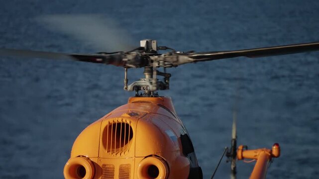 Close-up of an orange helicopter s spinning main rotor and upper fuselage on a maritime helideck, with motion blur revealing the swashplate, hub, and mechanical details.