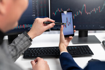 Cropped pointing hand with pen in businessman, checking with holding smartphone by woman investor online website program application with dynamic stock market at home office at back side. Infobahn.