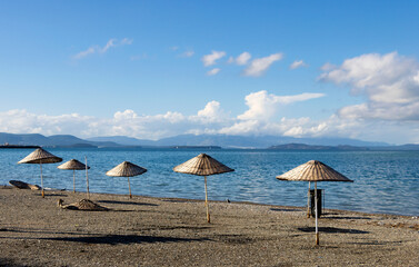 Row of Traditional Wicker Beach Umbrellas on a Quiet Pebble Beach, Overlooking a Calm Blue Sea and Distant Mountains Under a Bright Cloudy Sky.