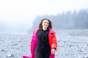 Smiling woman in a bright pink jacket walks joyfully along a rocky beach, surrounded by misty trees and a serene atmosphere, embodying freedom and adventure