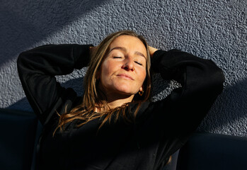 Woman with long hair relaxing against a textured wall, enjoying sunlight, with eyes closed, conveying a sense of peace and tranquility in a serene environment