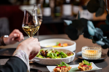 Close-up of a person holding a wine glass filled with white wine at a dining table with various gourmet dishes like salads and appetizers in a restaurant setting