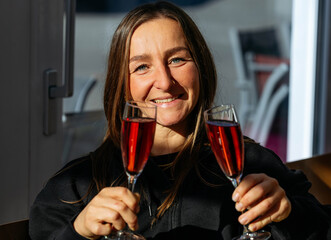 Smiling woman holding two glasses of sparkling beverage, illuminated by natural light, showcasing a joyful celebration moment with vibrant colors and festive atmosphere