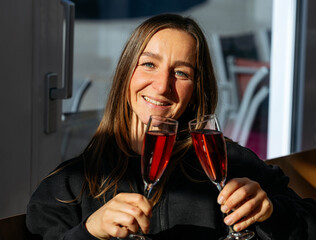 Woman with long hair, smiling brightly, holds two glasses of sparkling beverage, illuminated by warm sunlight, celebrating a joyful moment with friends in a cozy indoor setting