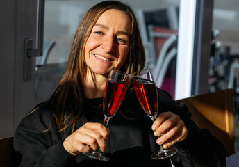 Smiling woman with long hair holds two glasses of sparkling red beverage, celebrating joyfully in a bright indoor setting, capturing a festive moment of happiness and togetherness