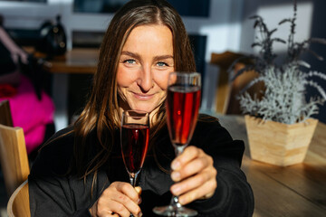Smiling woman with long hair holds two glasses of sparkling beverage, celebrating joyfully in a bright, modern cafe with warm sunlight illuminating the scene