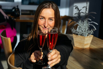 Woman with long brown hair is joyfully toasting with two glasses of sparkling beverage, surrounded by a cozy cafe atmosphere, showcasing celebration and happiness