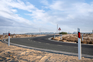Dukhan, Qatar - November 25, 2025: A deserted asphalt road curving through a dry desert landscape. Road signs warn of an upcoming bend