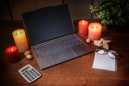 Laptop with empty screen, calculator and a notepad with glasses between candles and Christmas decoration on a dark brown wooden desk, online shopping, closed holiday business