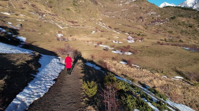 Side scrolling aerial view of a woman in a red coat walking through the countryside in winter with snowy mountains in the background