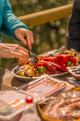 A woman cuts roasted vegetables, eggplants and red peppers with a knife and fork on a wooden table full of trays of meat and sauces to roast them on an outdoor barbecue.