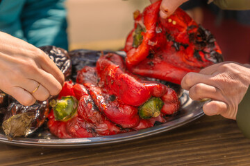 Young women's hands peeling roasted peppers and eggplants on a tray after they've been grilled on an outdoor barbecue.