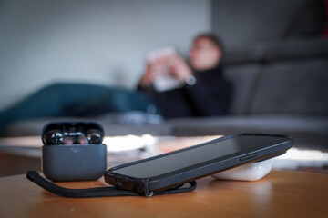 Smartphone and headphones set aside in the foreground while a man reads a book in the background. Concept of digital detox, disconnecting from technology, and returning to analog leisure