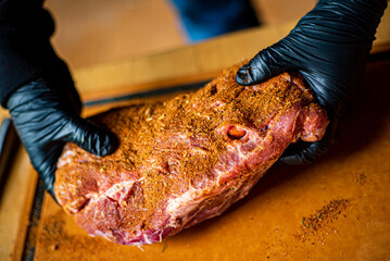 Hands wearing gloves hold a seasoned cut of meat, ready for grilling outside in warm weather