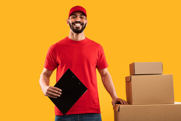 A delivery person is smiling and holding a clipboard in one hand. Several cardboard boxes are stacked next to him. The bright yellow background adds to the cheerful atmosphere.