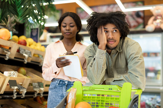 In a grocery store, a woman is taking notes on a clipboard while the man beside her leans on a shopping cart, showing visible frustration. Fresh produce is displayed nearby.