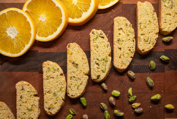 Biscotti cookies arranged in a diagonal line on a cutting board, with orange slices above and pistachio nuts below