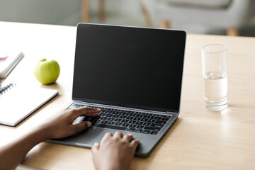 In a cozy home setting, an african american girl studies by typing on a laptop with a blank screen. She focuses on her homework while surrounded by a notebook, an apple, and a glass of water.