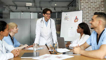 Smiling man chief physician making presentation in front of his male and female international colleagues during morning brainstorming at clinic, panorama. Medical conference, training concept