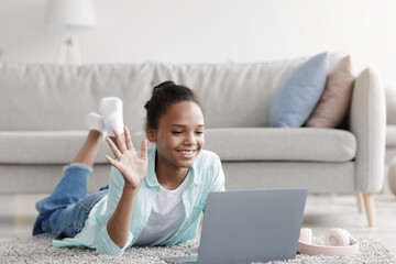 A cheerful young afro american girl lies on the floor in her living room, smiling as she waves her hand to the webcam on her laptop. She enjoys an online chat while staying safe during the pandemic.
