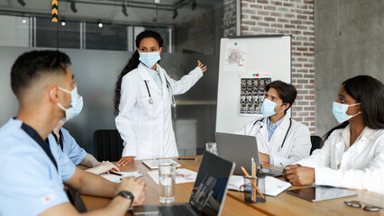 Multiracial group of men and women doctors in protective face masks having medical conference at clinic, physicians attending team-building, listening to middle-eastern woman chief doctor